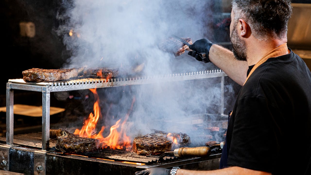Chef grilling steaks over open flame with smoke indoors, showcasing culinary expertise.